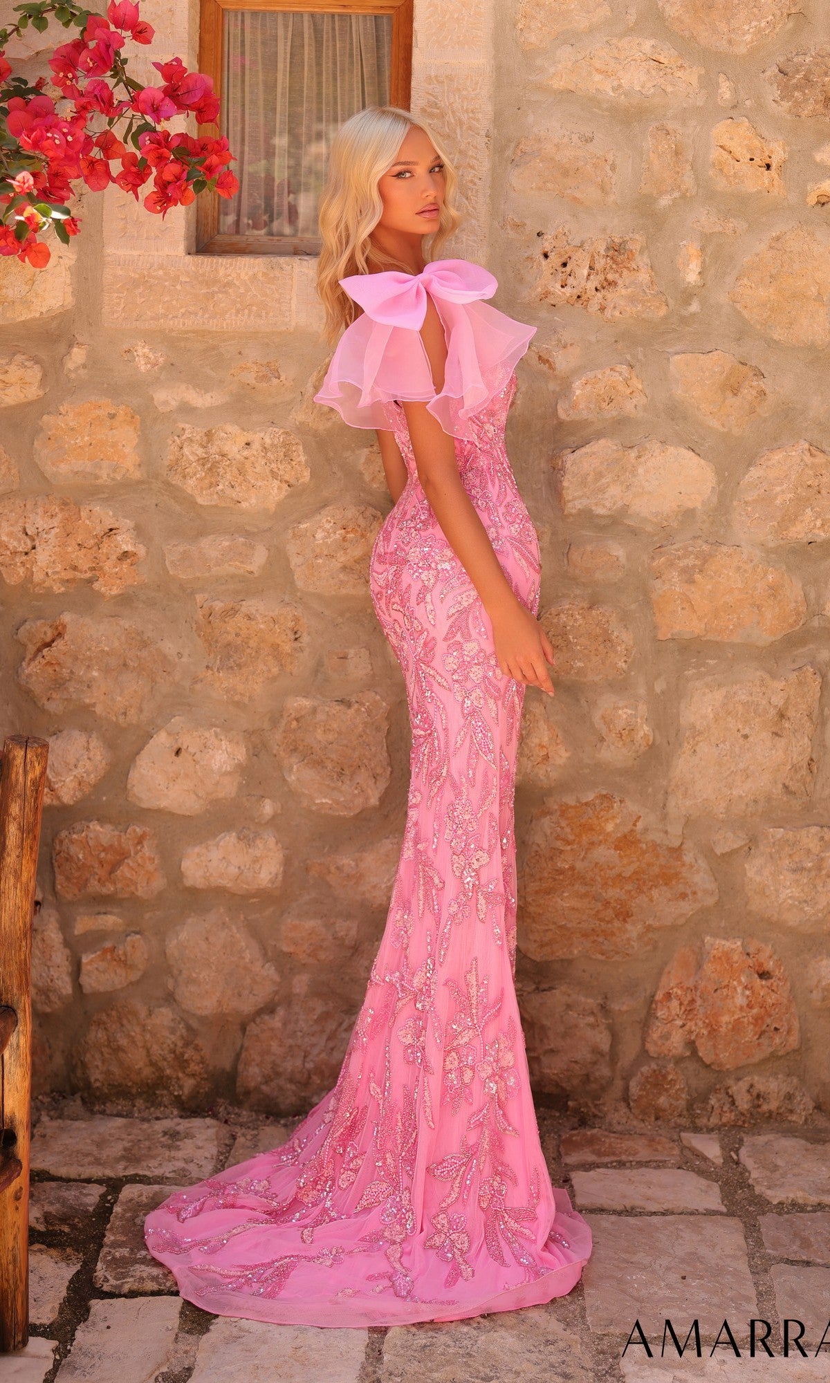 A woman in the Amarra 89175 long prom dress—a fitted, embellished pink one-shoulder gown with a large bow—stands against a stone wall beneath red flowers.